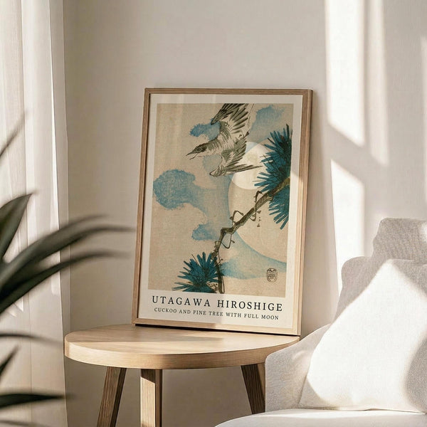 May include: Framed Japanese woodblock print reproduction featuring a cuckoo bird in flight with spread wings against blue clouds and a full moon, with pine tree branches visible. The artwork has muted earth tones and traditional ukiyo-e style. Displayed on a round wooden side table next to a beige upholstered chair, with plant leaves and natural window light creating shadows on white walls.