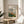May include: Framed art print displaying an impressionist landscape painting with green foliage, orange and yellow buildings, and blue sky. The poster features "Pierre Auguste Renoir" text and "view from Montmartre 1892" caption. Shown in thin wooden frame on round wooden side table next to cream upholstered chair, with plant shadows on white wall and natural sunlight streaming across the scene.