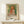 May include: Framed exhibition poster featuring a Renoir painting of a young girl with curly reddish hair wearing a white dress with orange vest, displayed on a light wood console table next to a small white wrapped gift with twine and evergreen sprig against a cream wall.