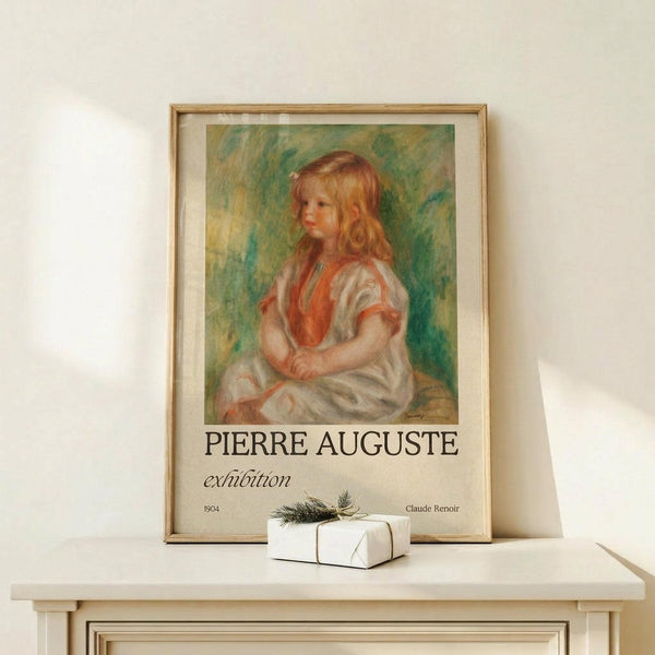 May include: Framed exhibition poster featuring a Renoir painting of a young girl with curly reddish hair wearing a white dress with orange vest, displayed on a light wood console table next to a small white wrapped gift with twine and evergreen sprig against a cream wall.