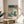 May include: Framed Japanese woodblock print displayed on round wooden side table next to beige upholstered chair. The artwork shows a snowy winter scene with people carrying umbrellas on a ferry crossing blue water, traditional buildings and trees in background under starry teal sky. Natural lighting creates soft shadows across white walls with plant visible in corner.