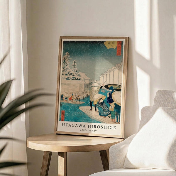 May include: Framed Japanese woodblock print displayed on round wooden side table next to beige upholstered chair. The artwork shows a snowy winter scene with people carrying umbrellas on a ferry crossing blue water, traditional buildings and trees in background under starry teal sky. Natural lighting creates soft shadows across white walls with plant visible in corner.