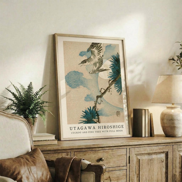 May include: Framed Japanese woodblock print featuring a flying bird with spread wings above pine branches and blue clouds against a beige background, displayed on a rustic wooden sideboard alongside fern plant, ceramic vase, books, and table lamp in a cozy living room setting with upholstered chair.