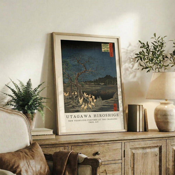 May include: Framed Japanese woodblock print reproduction featuring foxfires gathered beneath a bare tree at night, with deep blue sky and traditional red text cartouche. Light wood frame displayed on rustic wooden sideboard alongside fern plant, pottery vase, books, and cream upholstered chair with brown throw blanket in neutral-toned living room setting.