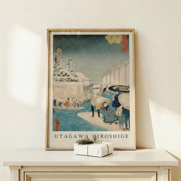 May include: Framed Japanese woodblock print featuring people with umbrellas crossing a snowy waterfront scene in traditional ukiyo-e style, displayed on light wood furniture with small wrapped gift, showing teal water, falling snow, and winter landscape with buildings and trees in muted colors