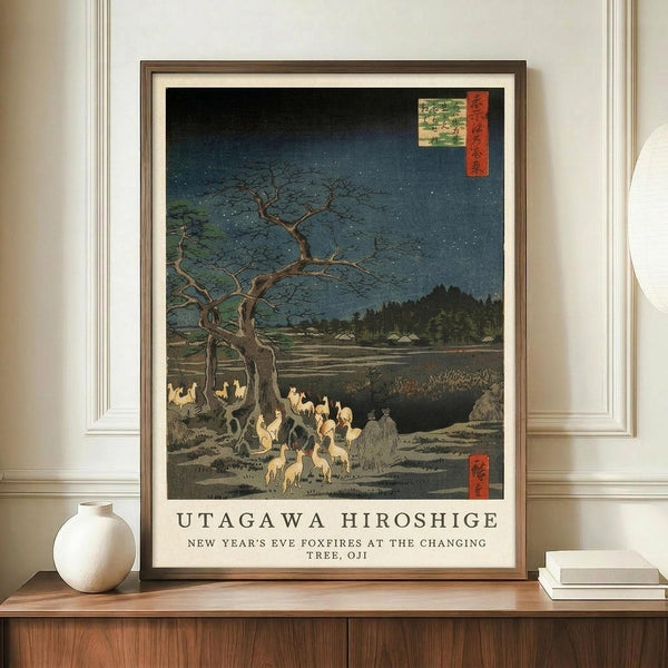 May include: A framed Japanese woodblock print reproduction displayed on a wooden sideboard next to a white ceramic vase and stacked books. The artwork depicts foxes with glowing flames gathering under a bare tree at night, with mountains and water in the background under a starry blue sky. The print features traditional ukiyo-e style with muted earth tones and red cartouche elements, housed in a brown wooden frame against white wainscoted walls.