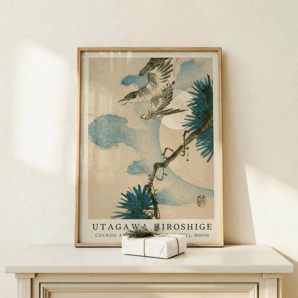 May include: Framed Japanese woodblock print featuring a cuckoo bird in flight against blue clouds and Mount Fuji, with pine branches in the foreground. The artwork displays traditional ukiyo-e style with muted earth tones and blue accents. Light wood frame sits on pale wooden furniture beside a small wrapped gift with greenery. Clean white wall background creates minimalist display setting.