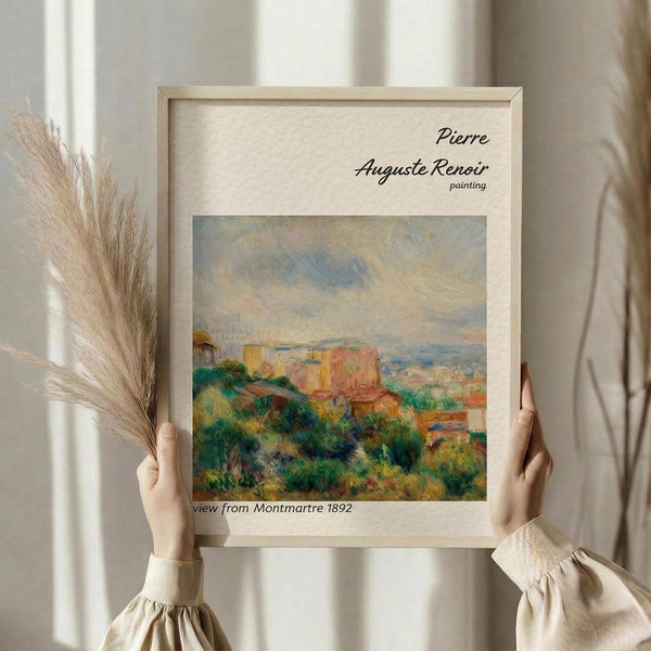 May include: Hands holding a framed print of a Renoir landscape painting showing colorful buildings and lush green vegetation from an elevated viewpoint, with dried pampas grass visible to the left, against white curtains and neutral background