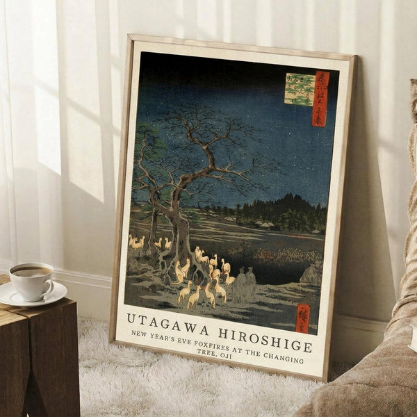 May include: A framed Japanese woodblock print reproduction leaning against a white wall on a textured white rug. The artwork shows foxes with glowing flames gathered beneath a bare tree under a starry night sky, featuring deep blue and green tones with traditional red text cartouches. A wooden side table with coffee cup and beige throw blanket are visible nearby.