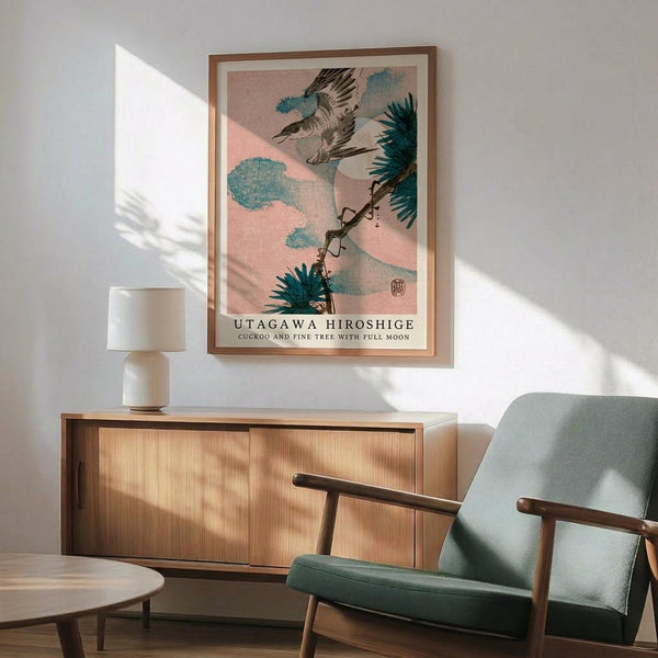 May include: Japanese woodblock print poster in wooden frame showing cuckoo bird flying near pine tree with full moon and turquoise clouds on pink sky background. Displayed above modern wooden credenza with white ceramic lamp in minimalist room with sage green mid-century armchair and natural lighting casting geometric shadows on white wall.