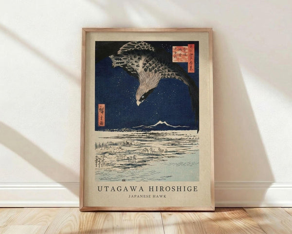 May include: Framed vintage Japanese woodblock print featuring a large hawk with detailed feathers flying against a deep blue night sky with white dots representing snow or stars. Below shows a winter landscape with Mount Fuji in the distance and scattered buildings across snowy fields. The artwork has traditional red Japanese text cartouches in the upper corners and sits in a light wooden frame against white walls on light wooden flooring with natural window light casting shadows.