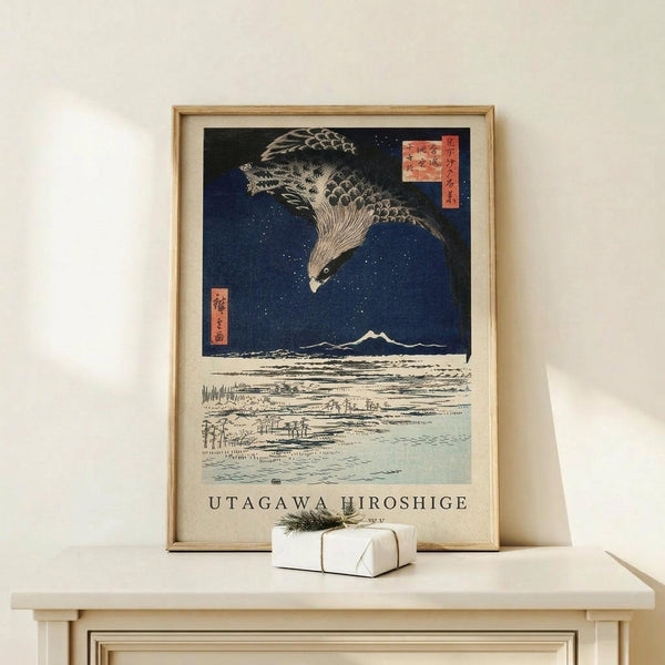 May include: Framed Japanese woodblock print featuring a large hawk with detailed feathers flying against a deep blue starry night sky above a snowy landscape with Mount Fuji in the distance. The artwork has traditional red cartouches with Japanese text and "UTAGAWA HIROSHIGE" printed below. Displayed on a light wood console table with a small white wrapped gift beside it against a cream wall.