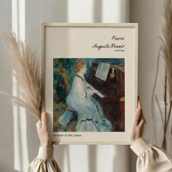 May include: hands holding a framed art print of Renoir's impressionist painting showing a woman in a white dress playing piano, with dried pampas grass stems, against white curtained background in natural lighting
