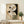 May include: Abstract geometric poster in wooden frame featuring curved shapes in beige and black, linear grid patterns, and circular elements, displayed on light wood console table with folded cream blanket and small potted evergreen plant in brown paper bag planter against white wall