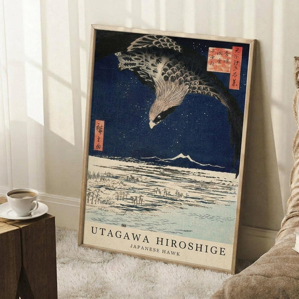 May include: Framed vintage Japanese woodblock print featuring a large hawk with detailed feathers flying over a snowy landscape with mountains under a deep blue starry sky. Print has traditional red text cartouches and cream border with English text. Wooden frame leans against white wall next to wooden side table with coffee cup and beige throw blanket on white textured rug.