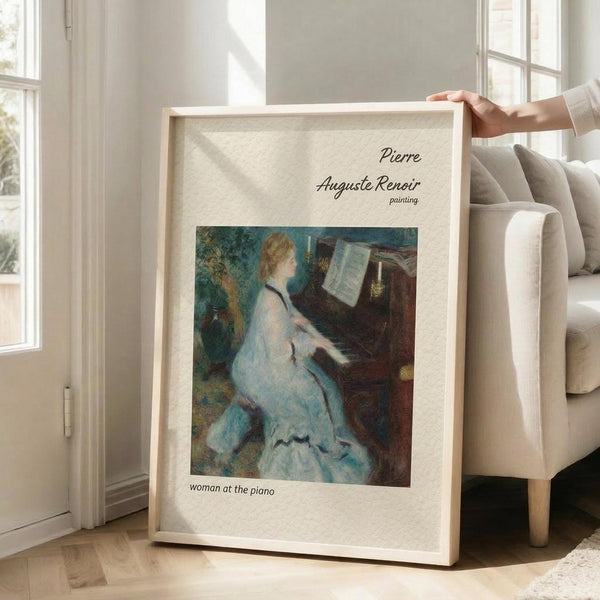 May include: Person's hands holding a framed poster print featuring Renoir's impressionist painting of a woman in a flowing white dress playing piano. The artwork shows soft blues and earth tones in Renoir's characteristic style. The print has a cream-colored mat with "Pierre Auguste Renoir painting" and "woman at the piano" text, set in a light wood frame. Scene shows bright modern living room with beige sofa, large windows, and natural lighting.