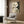 May include: A modernist abstract poster with geometric shapes in cream, black, and gold tones displayed on a wood-paneled wall above a beige upholstered armchair with geometric throw pillow. The artwork features overlapping circles, grid patterns, and linear elements in a neutral color palette. Scene includes a brass floor lamp and side table in a contemporary living room setting with natural lighting from sheer curtained windows.