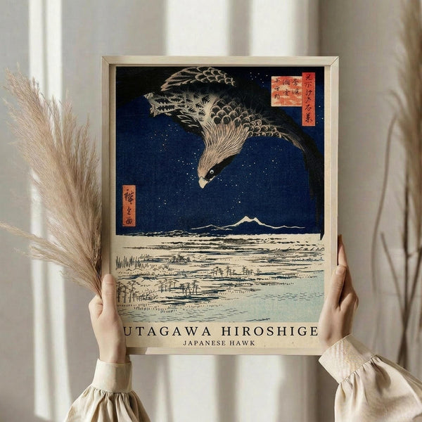 May include: Person in cream blouse holding framed Japanese woodblock print featuring large hawk with detailed feathers soaring over snowy landscape with Mount Fuji in background against deep blue starry sky. Print has traditional red text cartouches and cream mat border. Person also holds dried pampas grass stems. Neutral curtained background.