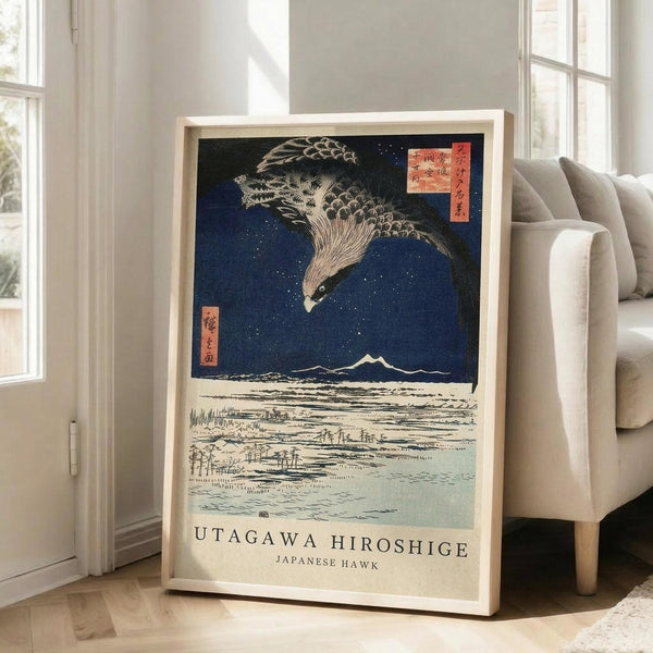 May include: Framed Japanese woodblock print featuring a large hawk with detailed feathers soaring above a snowy landscape with Mount Fuji in the distance under a deep blue starry sky. Red Japanese text cartouches visible in upper right corner. Print displayed in light wood frame leaning against white wall next to beige upholstered sofa in bright, naturally lit room with large windows.