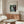 May include: Framed art print of Renoir's "Woman at the Piano" showing a woman in a white dress playing piano, displayed above a wooden sideboard with sliding doors in a modern living room with cream walls, white curved chair, glass oval coffee table, and brass floor lamp with circular mirror detail.