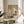 May include: Japanese-inspired artwork in wooden frame on round wooden table next to beige upholstered chair. Art features geometric composition with golden yellow rectangle, coral red circle, black linear elements, and delicate pine branch illustrations in muted earth tones. Natural lighting creates soft shadows across white wall background with plant leaves visible at left edge.