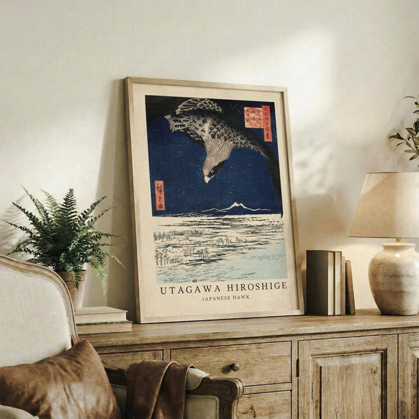 May include: Framed Japanese woodblock print featuring a detailed hawk in flight against deep blue starry sky above snowy landscape, displayed on rustic wooden sideboard alongside fern plant, books, and ceramic vase in neutral-toned living room setting with cream upholstered furniture and warm natural lighting.