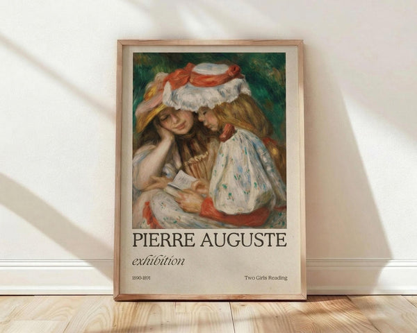May include: Framed exhibition poster showing an impressionist painting of two young women reading together under a shared hat, rendered in soft pastels of green, white, and coral. The wooden frame sits against a white wall on light wood flooring with natural window shadows creating diagonal light patterns across the scene.