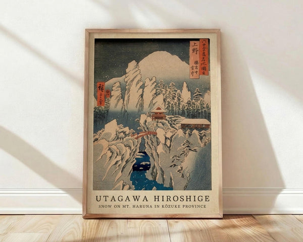 May include: Framed Japanese woodblock print reproduction showing snow-covered mountains, traditional temple buildings, and deep blue water flowing through rocky gorge. Features muted earth tones with red text cartouches in upper corners against dark sky with falling snow. Natural wood frame displayed on light wooden floor against white wall with soft window light creating diagonal shadows.