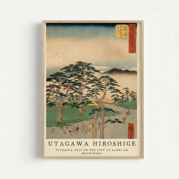 Japanese woodblock print in light wood frame showing Mount Fuji in the distance with pine trees in the foreground and small figures of travelers on a path. The artwork features soft blues, greens, and earth tones with traditional red text cartouches in the upper corners. Text below reads "UTAGAWA HIROSHIGE" and "FUJISAWA: FUJI ON THE LEFT AT NANKI NO MATSUBARA" against a cream background.