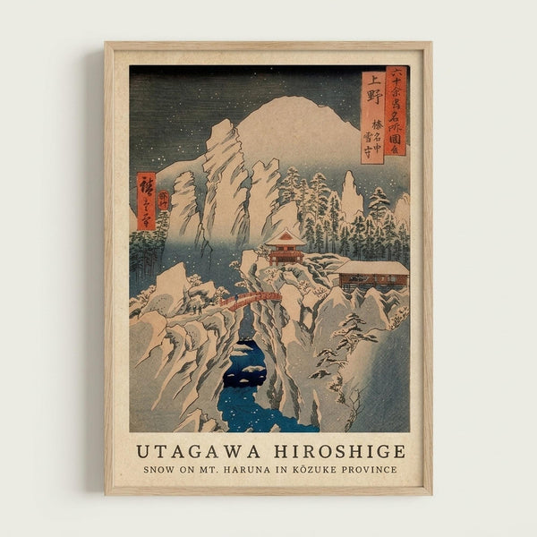 A framed Japanese woodblock print showing a snowy winter mountain landscape with steep cliffs covered in white snow. A blue river winds through the rocky canyon below, while traditional buildings with red roofs nestle among snow-laden pine trees on the mountainsides. The dark sky is filled with falling snow depicted as small white dots. The artwork features muted colors - grays, blues, whites and touches of red - in the classic ukiyo-e style. Red text cartouches with Japanese characters appear i
