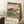 May include: Framed Japanese woodblock print reproduction leaning against white wall on textured rug. Artwork shows Mount Fuji in background with large pine trees, small figures walking along a path, in traditional ukiyo-e style with blue gradient sky and green landscape. Light wood frame, displayed next to wooden cube with white coffee cup and saucer, soft natural lighting from window.