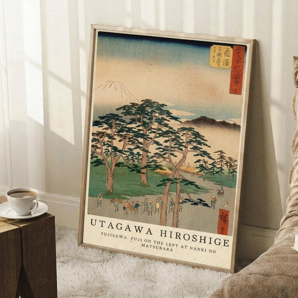 May include: Framed Japanese woodblock print reproduction leaning against white wall on textured rug. Artwork shows Mount Fuji in background with large pine trees, small figures walking along a path, in traditional ukiyo-e style with blue gradient sky and green landscape. Light wood frame, displayed next to wooden cube with white coffee cup and saucer, soft natural lighting from window.