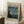 May include: Framed Van Gogh museum poster featuring The Starry Night painting with swirling blue sky, bright yellow stars, dark cypress tree, and village below. Light wood frame leans against white wall corner next to wooden side table with white coffee cup. Beige textured rug and tan blanket visible in cozy room setting.