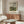 May include: Framed art print featuring an impressionist landscape painting with cloudy sky, river, and village buildings, displayed above a walnut wood credenza in a modern living room with white curved chair, glass oval coffee table, and minimalist decor including brass floor lamp and table lamp