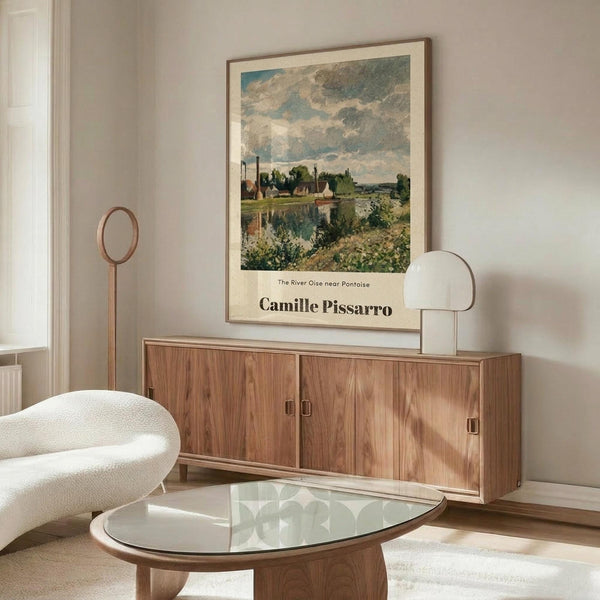 May include: Framed art print featuring an impressionist landscape painting with cloudy sky, river, and village buildings, displayed above a walnut wood credenza in a modern living room with white curved chair, glass oval coffee table, and minimalist decor including brass floor lamp and table lamp