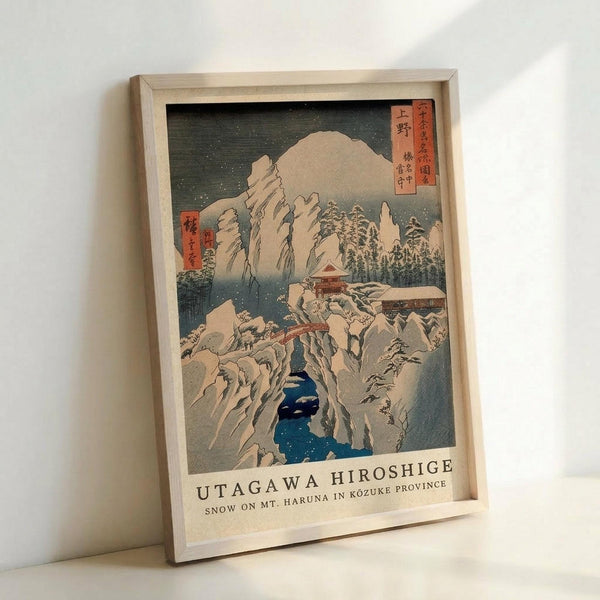 Framed Japanese woodblock print reproduction showing a snowy mountain landscape with dramatic rocky cliffs, traditional buildings nestled among snow-covered trees, and a deep blue stream or gorge cutting through white winter scenery. The artwork features muted colors with red text cartouches in the upper corners, displayed in a light wooden frame leaning against a white wall.