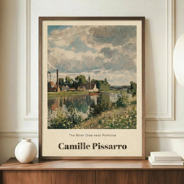 May include: Framed Impressionist-style landscape print showing a river scene with houses, trees, and cloudy sky in muted blues, greens, and earth tones. The artwork features Camille Pissarro's name at bottom. Print displayed on wooden credenza beside white ceramic vase and stacked books against cream-colored wall with white molding trim.
