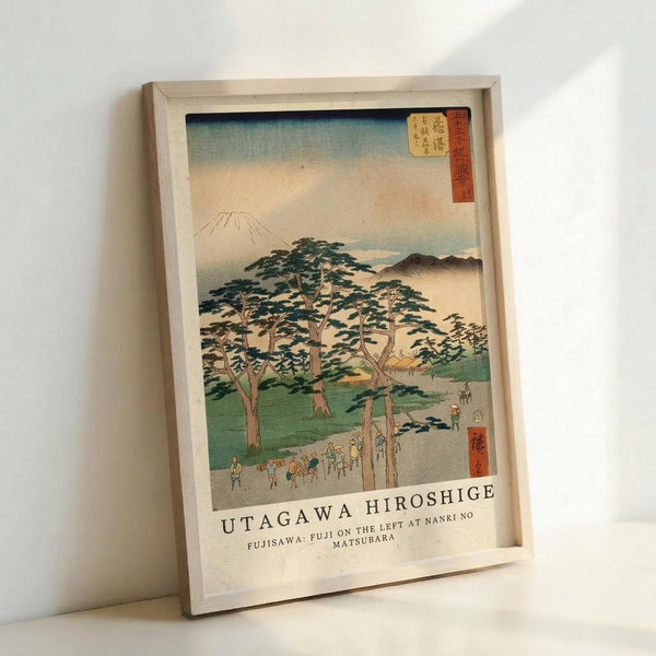 A framed Japanese woodblock print showing Mount Fuji in the distance with rolling hills and a grove of tall pine trees in the foreground. Small figures of travelers walk along a path beneath the trees. The artwork features soft blues, greens, and peachy tones with traditional red cartouches containing Japanese text. The print is displayed in a light wooden frame leaning against a white wall, with the artist name Utagawa Hiroshige and location details printed below the image.