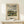 May include: Framed art print displaying an impressionist landscape painting with cloudy blue sky, riverside village scene with buildings and green foliage, displayed on light wood console table next to small white wrapped gift with green botanical accent against cream wall