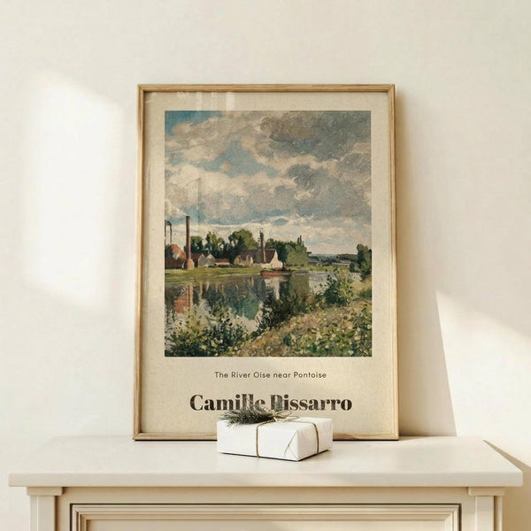 May include: Framed art print displaying an impressionist landscape painting with cloudy blue sky, riverside village scene with buildings and green foliage, displayed on light wood console table next to small white wrapped gift with green botanical accent against cream wall