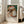 May include: Framed exhibition poster featuring Renoir's painting of two young girls in white dresses and bonnets reading together, displayed in bright modern living room with beige sofa, white walls, and large windows. Poster has cream matting in light wood frame, leaning against wall on hardwood floor.