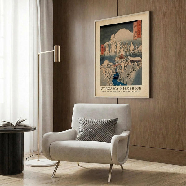 May include: Framed vintage Japanese woodblock print showing snow-covered mountain landscape with blue water and traditional buildings, displayed on wood-paneled wall above cream upholstered armchair with geometric patterned pillow. Modern brass floor lamp and dark side table with open book visible in contemporary living room setting with warm wood tones and neutral textiles.