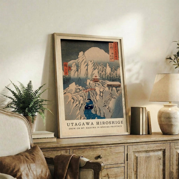 May include: Japanese woodblock print poster in light wood frame displayed on rustic wooden sideboard. Print shows snow-covered mountains, dark blue water, and small buildings in traditional ukiyo-e style with red text blocks. Scene includes cream sofa with brown pillow, green fern plant, stacked books, ceramic vase, and warm lighting against white wall.