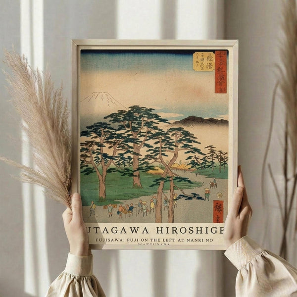 May include: Person in beige clothing holding framed Japanese woodblock print showing Mount Fuji in background with large pine trees in foreground, small figures walking on path, and traditional buildings. Print features soft blue sky, green landscape, and Japanese text in red cartouche. Light wood frame against white curtained window with dried wheat stalks visible on left side.