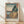 May include: Framed Japanese woodblock print poster showing coastal landscape with rocky cliffs, blue ocean waves, and distant mountains under pale sky. Print features traditional ukiyo-e art style with muted blues, greens, and earth tones. Wooden frame sits on light wood floor against white wall with natural window light casting shadows.