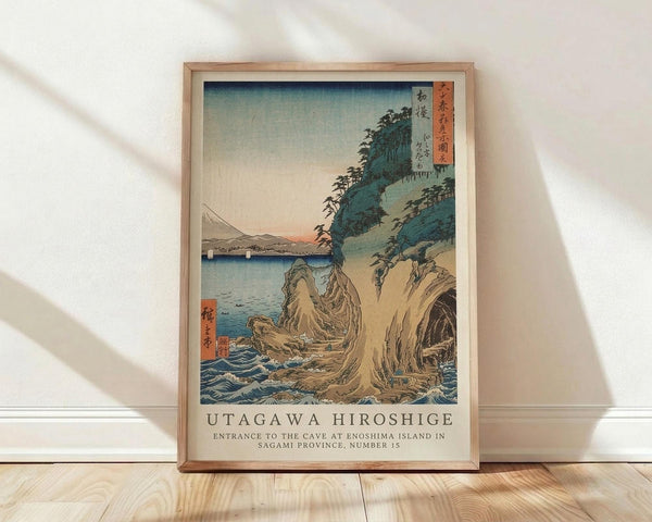 May include: Framed Japanese woodblock print poster showing coastal landscape with rocky cliffs, blue ocean waves, and distant mountains under pale sky. Print features traditional ukiyo-e art style with muted blues, greens, and earth tones. Wooden frame sits on light wood floor against white wall with natural window light casting shadows.