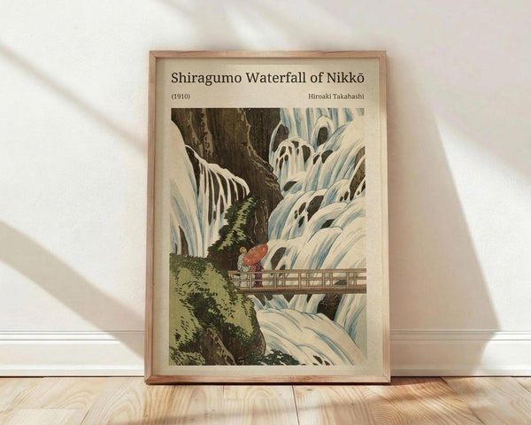 May include: A framed Japanese woodblock print reproduction displayed on light wooden flooring against a white wall with natural shadows. The artwork depicts a dramatic waterfall scene with cascading white and blue water, dark rocky cliffs, green vegetation, and a small figure with a red umbrella crossing a wooden bridge. The print is matted in cream and housed in a light wood frame.