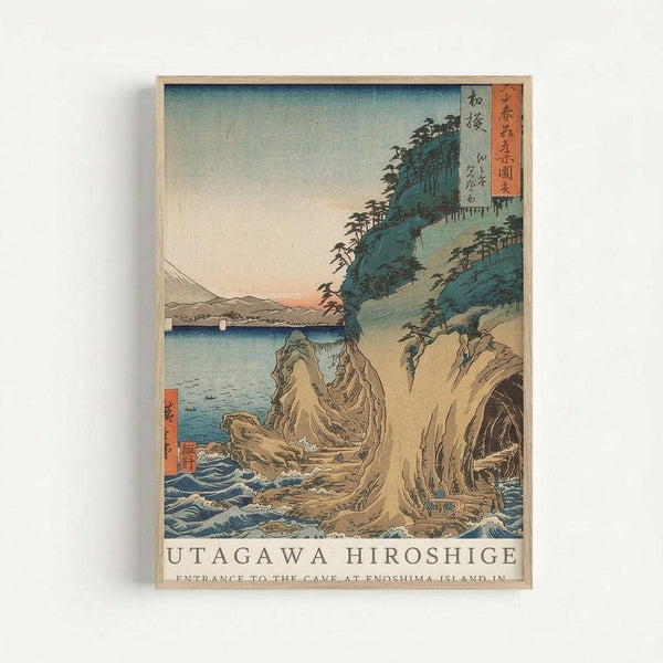 Japanese woodblock print depicting a dramatic coastal scene with towering rocky cliffs and crashing waves at Enoshima Island. The artwork features traditional ukiyo-e style with muted blue-green tones for the forested cliffs, warm peachy sky tones, and detailed wave patterns in the foreground. Red text cartouches with Japanese characters appear in the upper right and lower left corners. The print is displayed in a light wooden frame against a white wall, showing the classic horizontal format typ