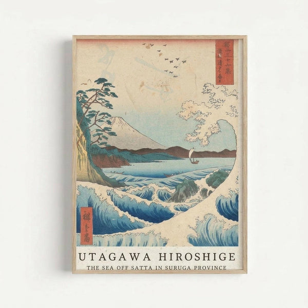Framed Japanese woodblock print reproduction showing dramatic ocean waves in the foreground with white foam caps and deep blue water. A lone pine tree bends on the left side, Mount Fuji appears in the background, and a small sailboat navigates between the waves. Flying birds dot the pale sky above rolling hills. Red text cartouches with Japanese characters appear in upper right and lower left corners. The artwork displays classic ukiyo-e style with soft earth tones and blues, mounted in a light