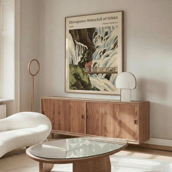 May include: Japanese woodblock print artwork depicting a dramatic waterfall with cascading white and blue water, wooden bridge, and green foliage, displayed in a thin wooden frame above a mid-century modern walnut credenza. The vintage-style poster is shown in a bright, minimalist living room with white walls, cream upholstered chair, glass coffee table, and modern table lamp.